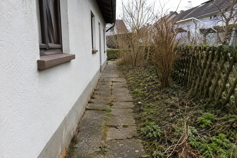 Alter Plattenweg an einer Hauswand mit schiefen, bemoosten Betonplatten